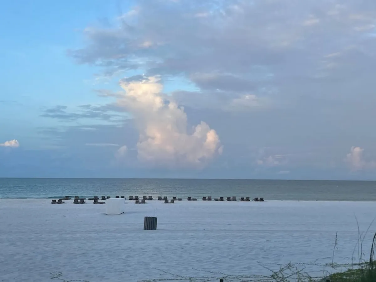 This image captures a beautiful stretch of white sandy beach lined with beach chairs facing the gentle waves of the ocean. The light, clouds, and footprints in the sand evoke a sense of peace and ease of direct beachfront access.