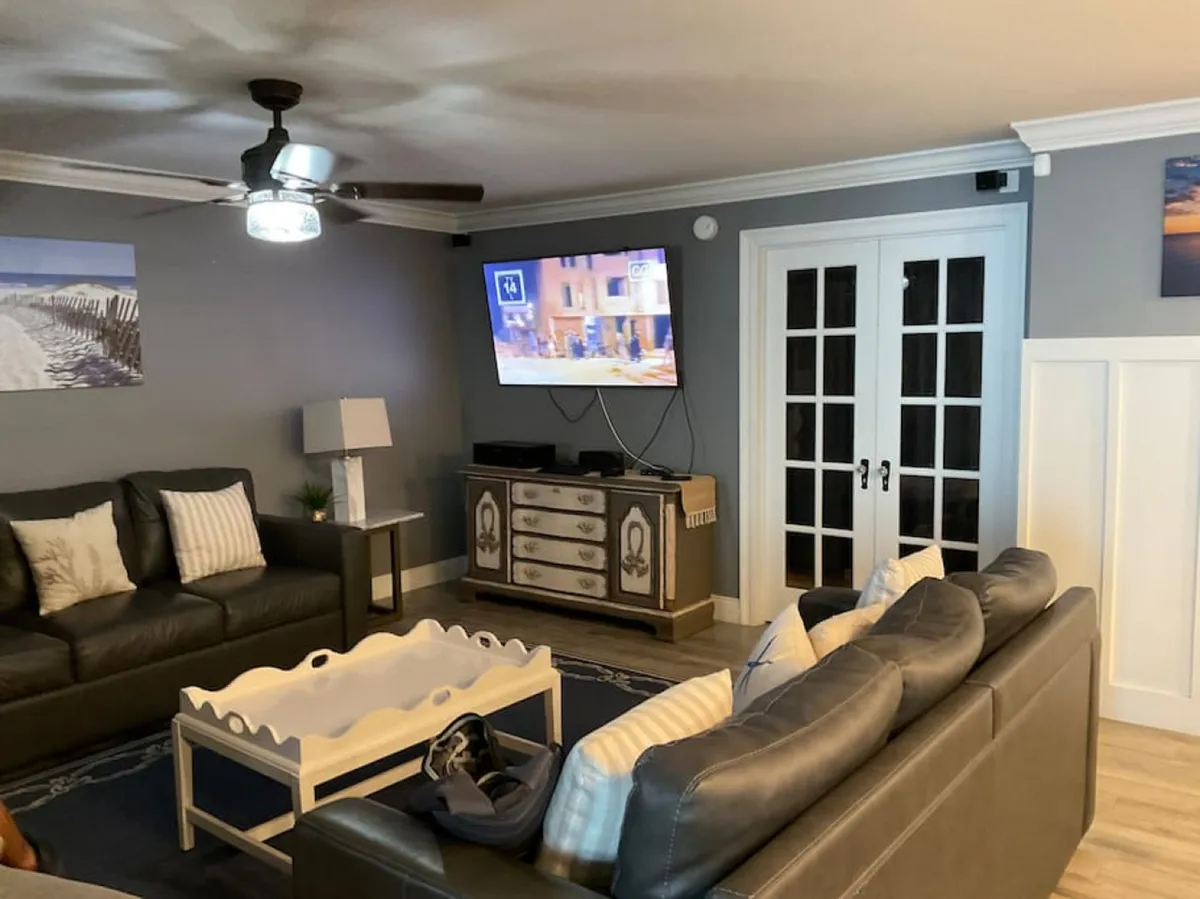 The image showcases a bright, open living area featuring two plush dark leather sofas adorned with striped and solid pillows, a charming white scalloped-edge coffee table at the center, a wall-mounted flat screen TV perfect for movie nights, and a decorative sideboard with drawers—all set in a relaxed, coastal-inspired space designed for gathering and unwinding after sun-filled beach days.