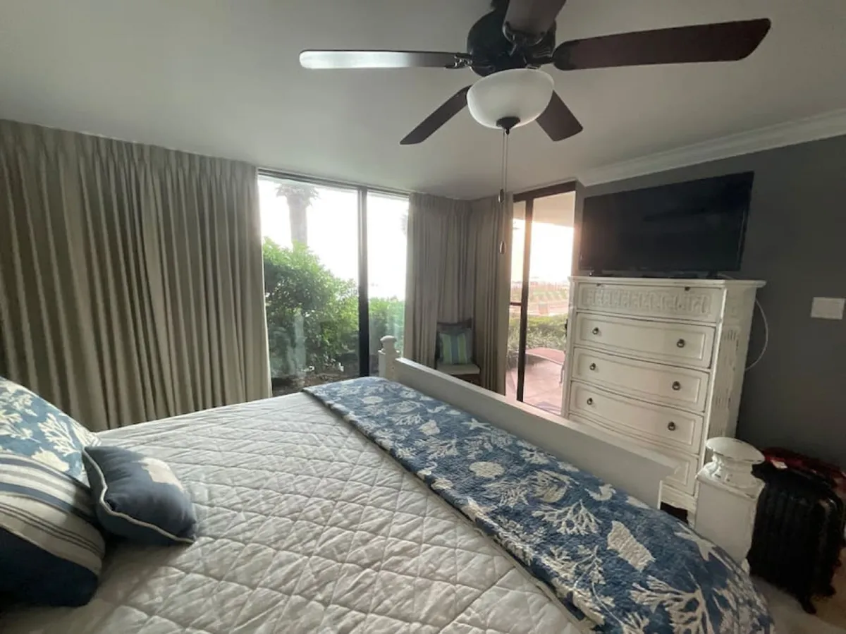 A spacious bedroom featuring a large bed with blue and white patterned bedding, a white dresser topped with a flat screen TV, and a sliding glass door leading to an outdoor patio. The ceiling fan with light fixture adds comfort, and the room is filled with natural light, creating a serene and restful atmosphere.