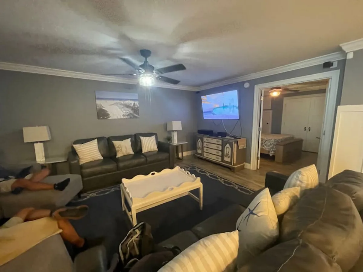 A spacious living area with large, dark leather sofas adorned with decorative pillows, a white coffee table, and a wall-mounted flat-screen TV. An open doorway reveals a bedroom beyond, suggesting a welcoming, communal space ideal for group gatherings, lounging, or shared activities.
