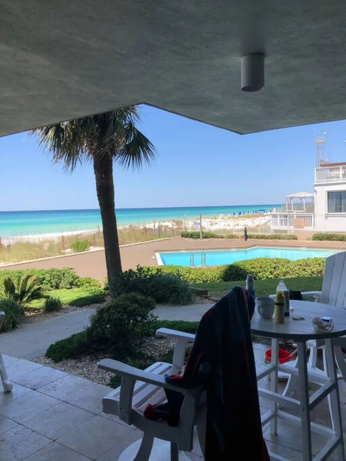 This scene features a turquoise ocean and sandy beach beside an outdoor swimming pool. A palm tree and a patio seating area with a table and chairs invite relaxation and outdoor meals, with the beach just steps away.
