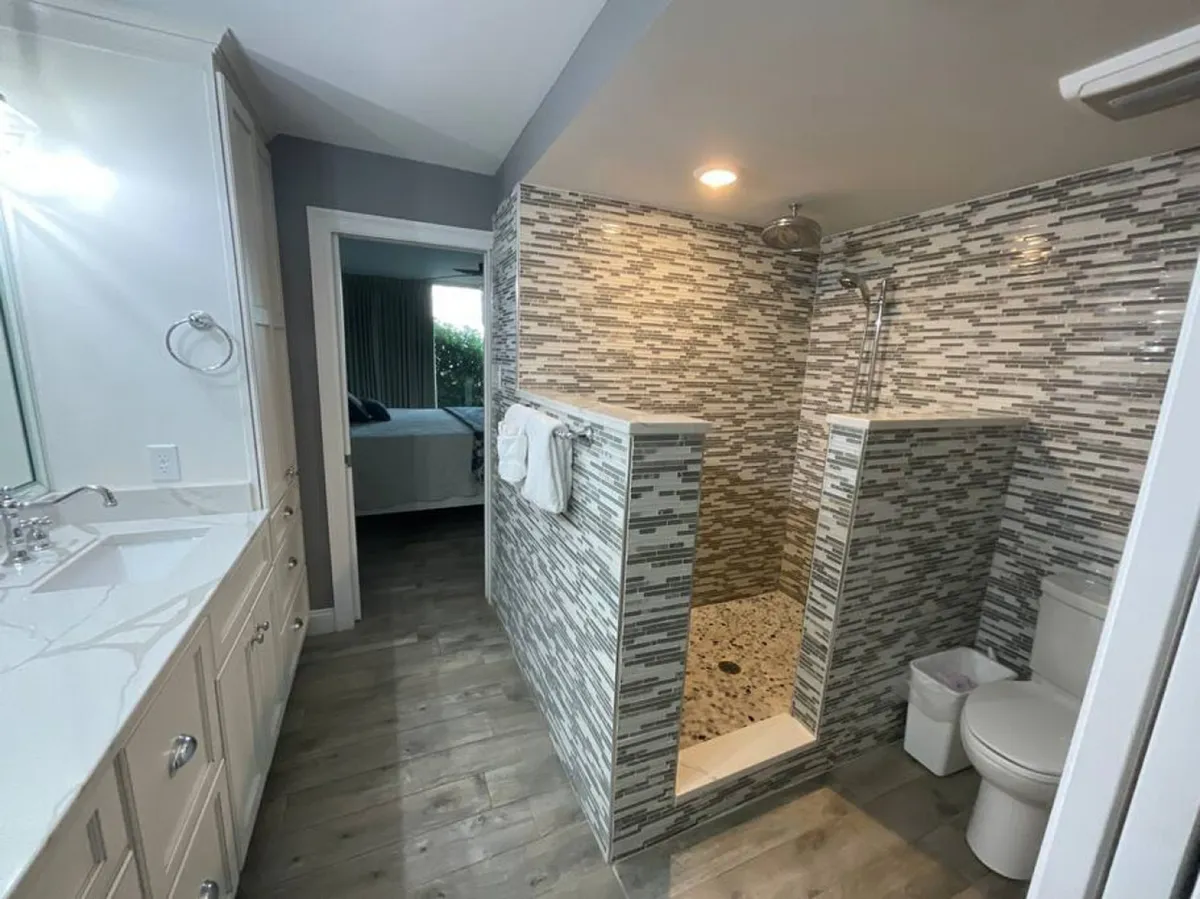 A modern bathroom with a walk-in shower featuring mosaic tile walls, a double sink vanity with a white countertop, and a view into an adjoining bedroom. The space is bright, fresh, and designed for comfort and convenience.