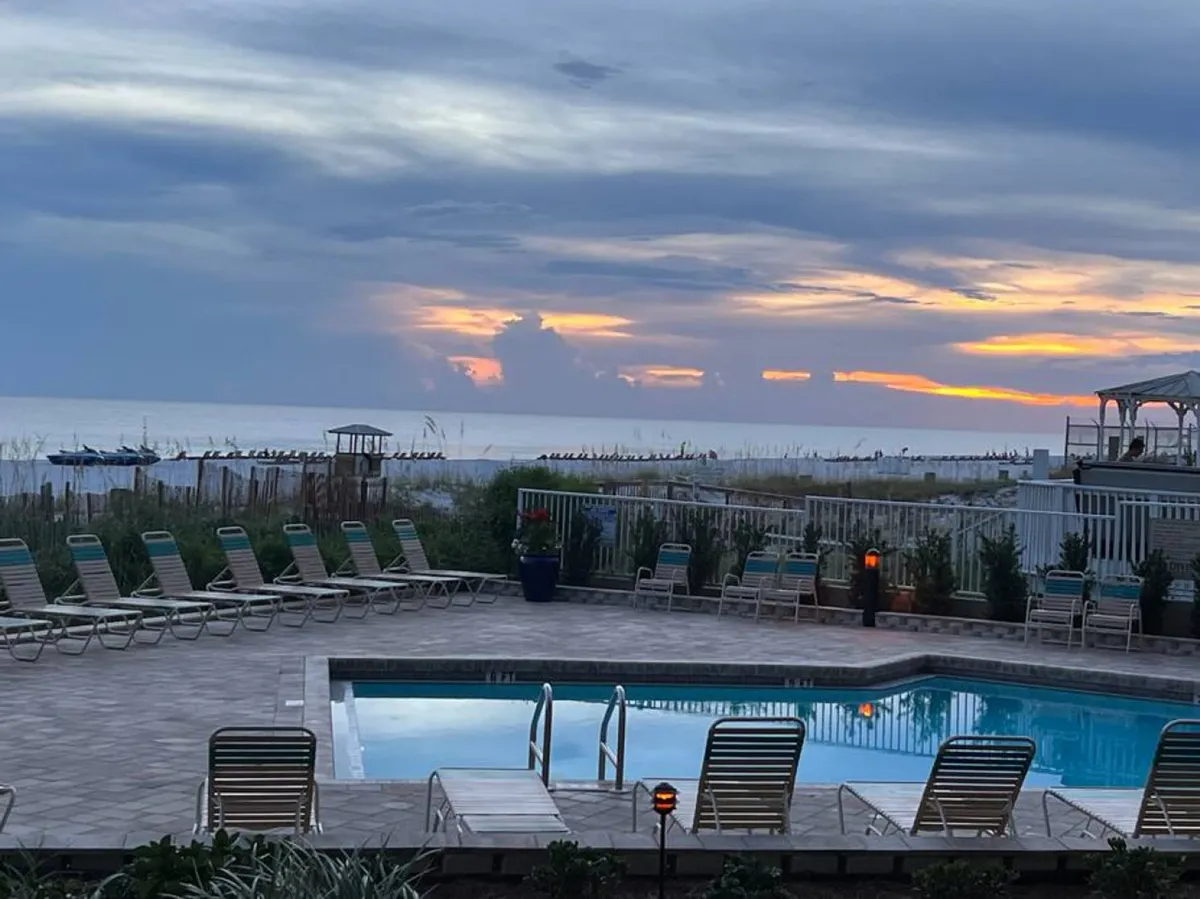 A swimming pool with clear water is surrounded by sun loungers, set against the backdrop of the ocean horizon and a colorful sunset sky. Dune grass and a fence separate the pool area from the beach, evoking a serene, coastal atmosphere ideal for unwinding after a meal.