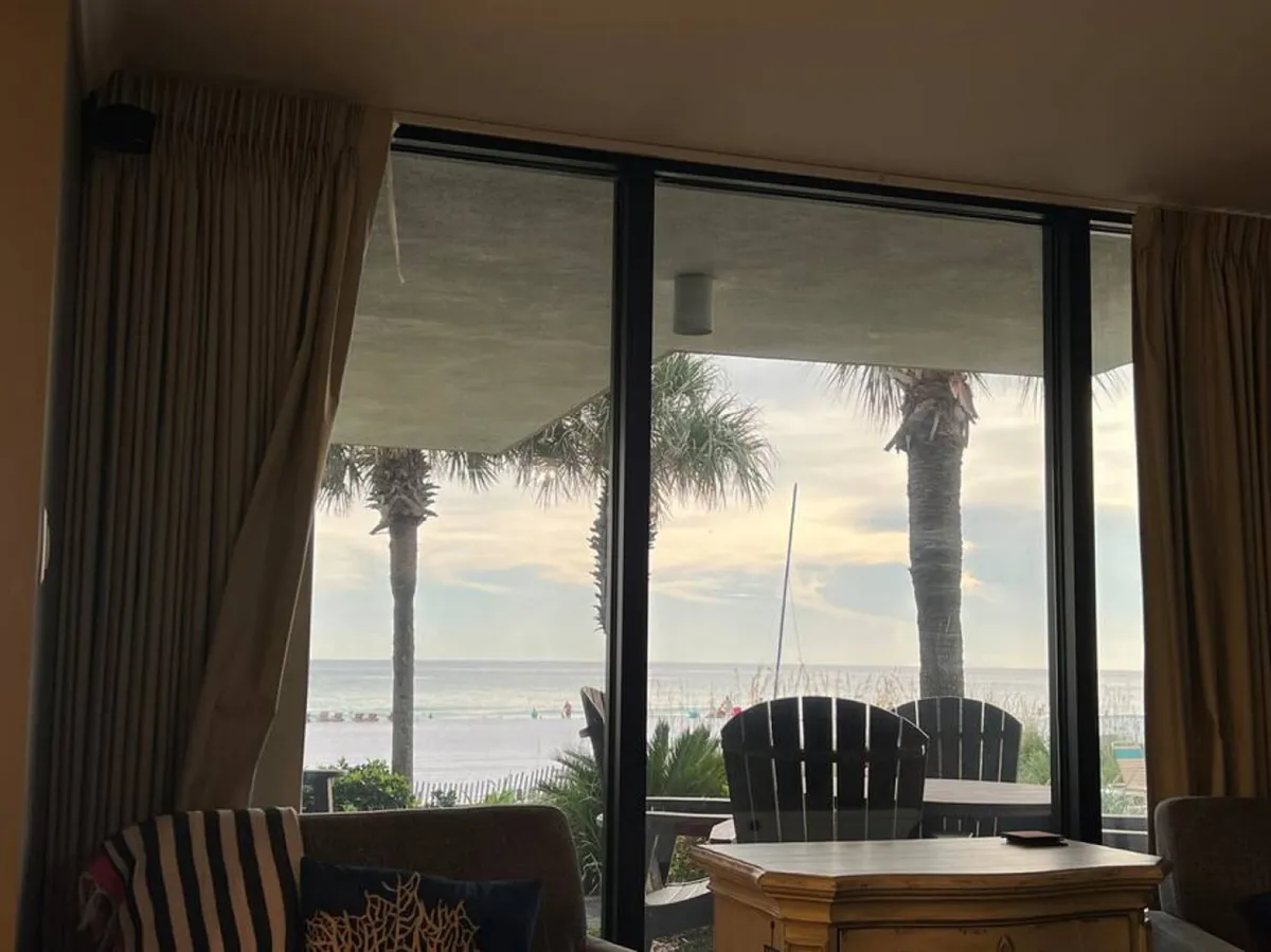 A welcoming living area with large sliding glass doors framing a lush beach view. Indoor wooden furniture is paired with outdoor patio seating, and palm trees are visible just outside, suggesting the ease of moving between interior relaxation and beachfront enjoyment.