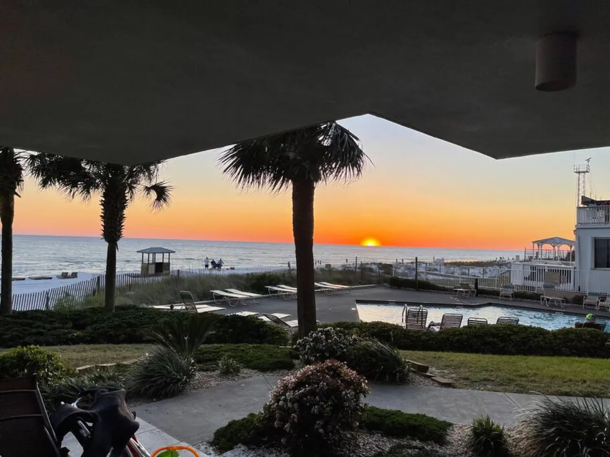 This image captures a stunning sunset over the ocean, with palm trees framing the view, a swimming pool in the foreground, and beach loungers set on the sandy shore. The scene showcases the seamless transition from poolside relaxation to beach exploration and evening gatherings.