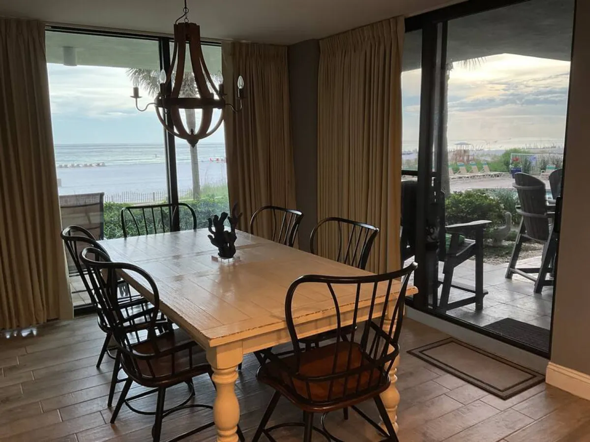 A spacious interior dining area featuring a large wooden table with black metal chairs. Panoramic glass doors open directly to a stunning ocean and beach view, seamlessly connecting the indoor space with the sandy exterior. The abundance of natural light and the clear sightline to the beach highlight the effortless indoor-outdoor flow.