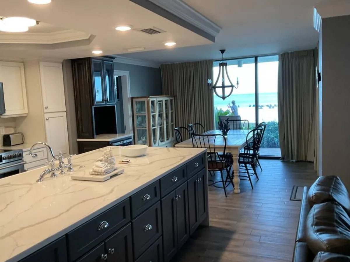 A bright, elegant open-plan kitchen and dining area with a large marble-topped island and a dining table set near sliding glass doors. The doors provide a direct view of the ocean and beach, reinforcing the flow from indoor family spaces to the outdoors.