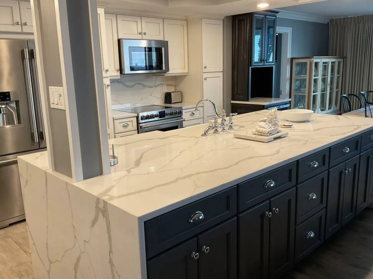 A spacious, modern kitchen featuring a large marble island with a built-in sink, stainless steel appliances including a refrigerator and oven, and sleek white cabinetry. The open layout seamlessly connects the kitchen to the adjacent dining area, allowing for easy interaction and entertaining.