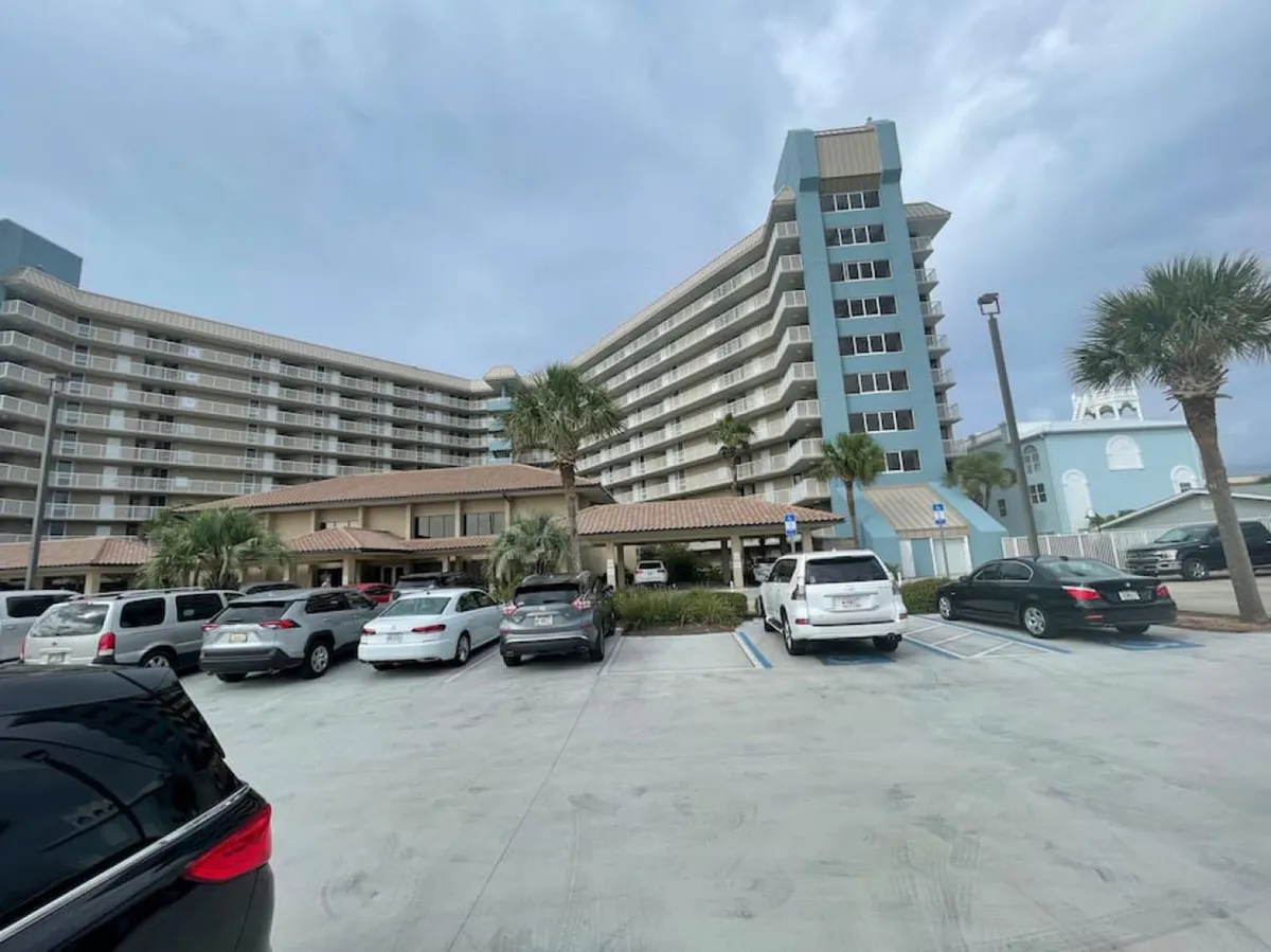 A large multi-story condo building with balconies, a covered entrance with a tiled roof, palm trees surrounding the property, and a parking lot filled with cars. The image captures the inviting and practical exterior, hinting at easy access to nearby amenities.