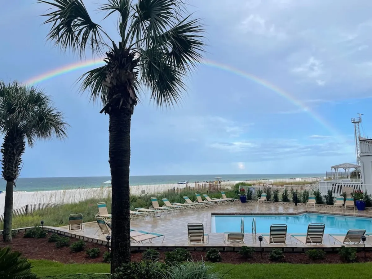 An exterior shot with a rainbow arching over the ocean, palm trees framing the pool area, and the oceanfront with white sand visible. Lounge chairs invite relaxation, and the view highlights both poolside and beachfront access.