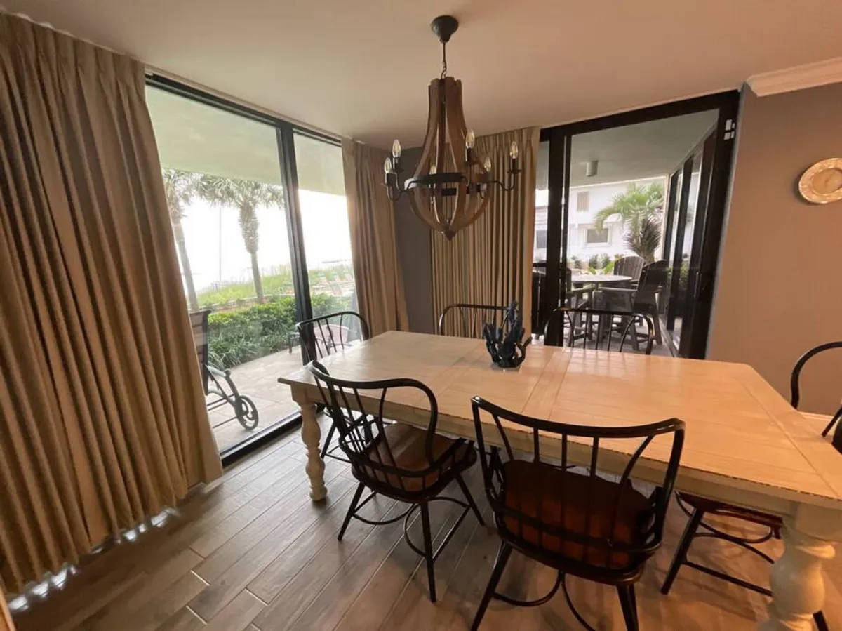 A bright dining area with a large white table surrounded by black metal chairs with cushions, illuminated by a wooden chandelier. Glass sliding doors provide an inviting outdoor view and fill the space with warm coastal light, creating a perfect setting for meals and laughter.