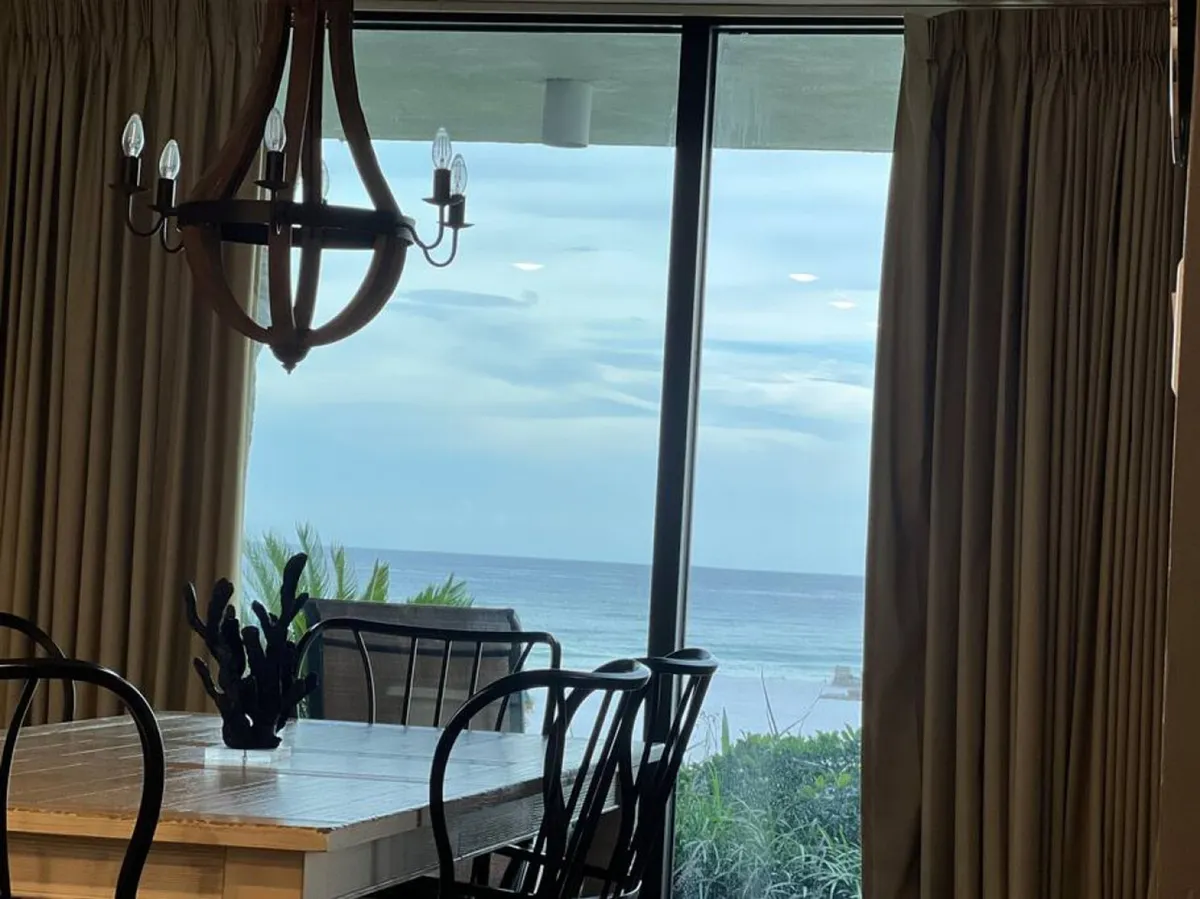 A large wooden dining table surrounded by black metal chairs, illuminated by a chandelier, with an ocean view visible through sliding glass doors. The inviting setting hints at easy indoor-outdoor living and relaxation after exploring the local area.