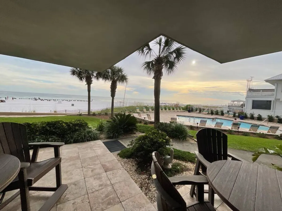 An ocean beach in the background, a swimming pool surrounded by lounge chairs, palm trees creating a tropical atmosphere, and a covered patio with wooden chairs and a round table. The space feels leisurely and inviting, blending indoor and outdoor relaxation.