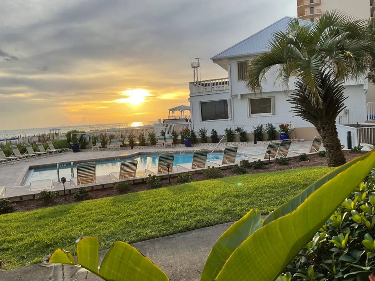 This exterior image features an inviting outdoor swimming pool surrounded by lounge chairs, lush tropical landscaping, and a white building with balconies overlooking the pool and beach. The sun is setting over the ocean horizon, casting a warm, golden glow across the entire scene.